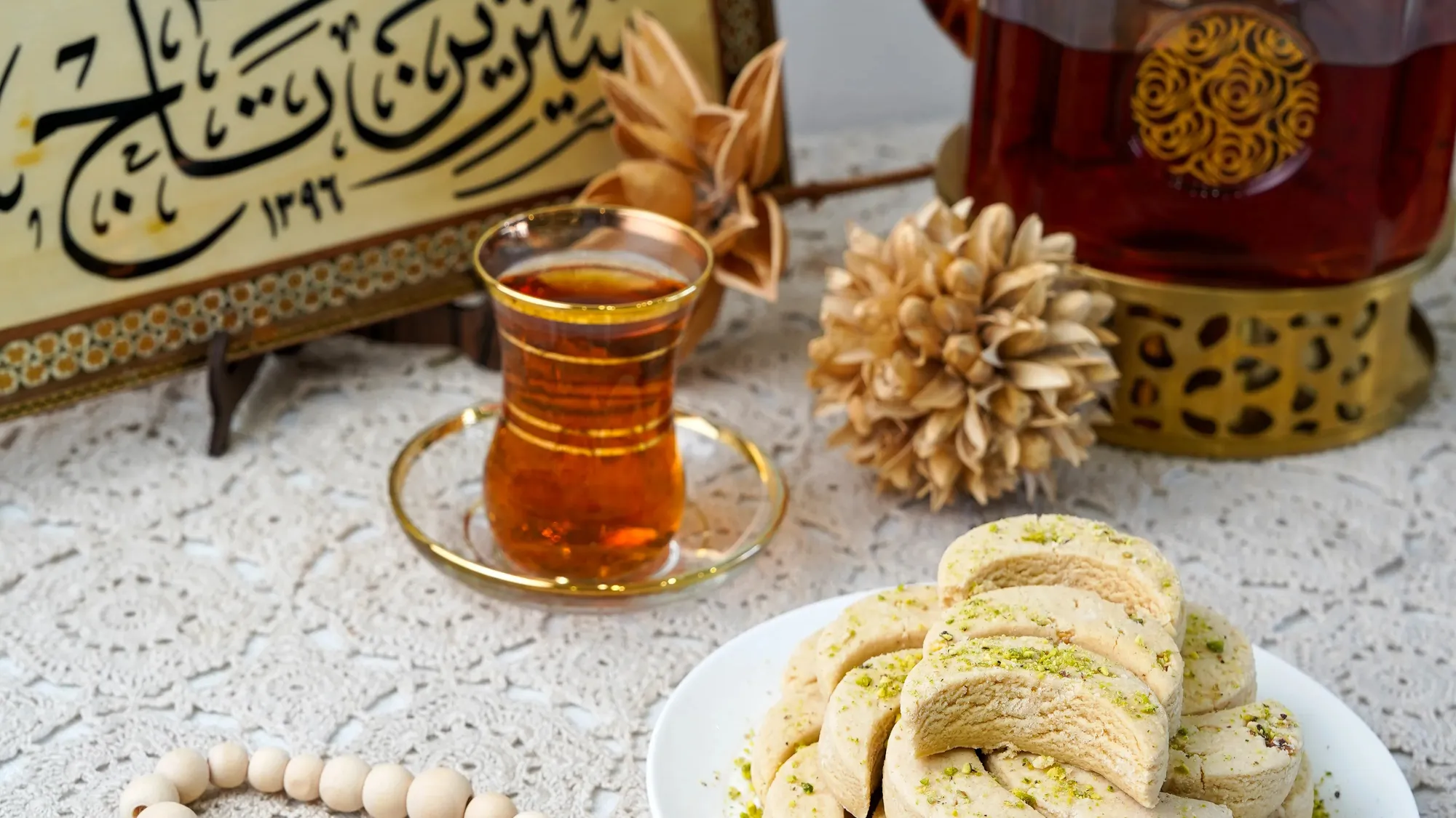 Persian tea in a gold-rimmed glass with pistachio sweets, in front of Sweet Crown's Shirin Taj calligraphy